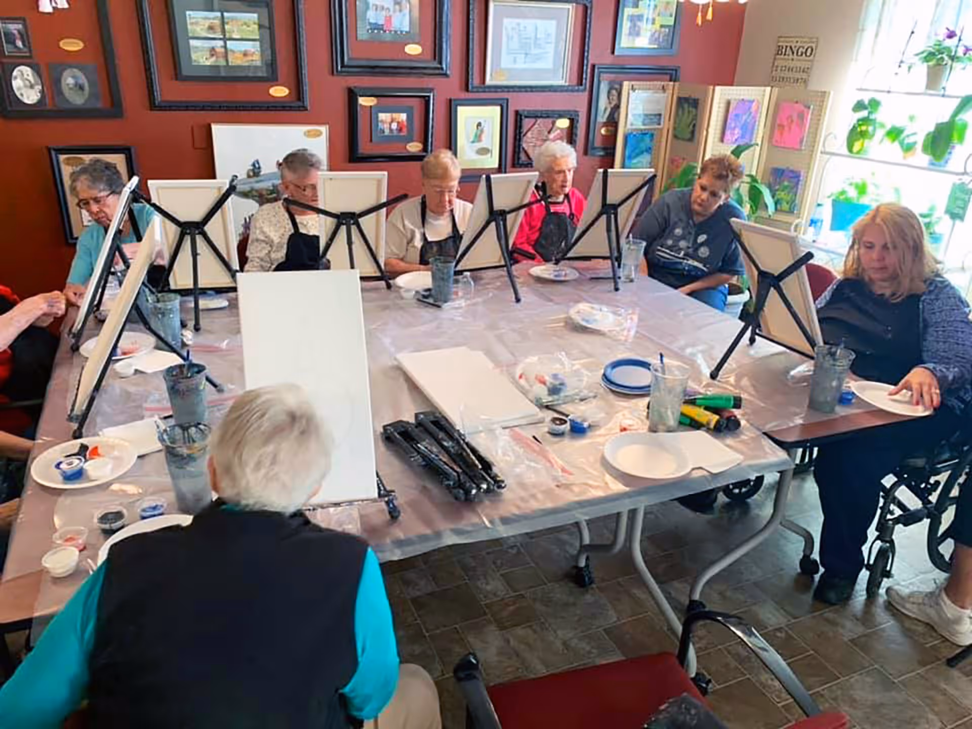 A group of elderly individuals seated around a large table engaged in a painting activity. Each person has a canvas on an easel in front of them, along with paint supplies and water cups. The room has framed pictures on the walls and a window with plants on the right side.