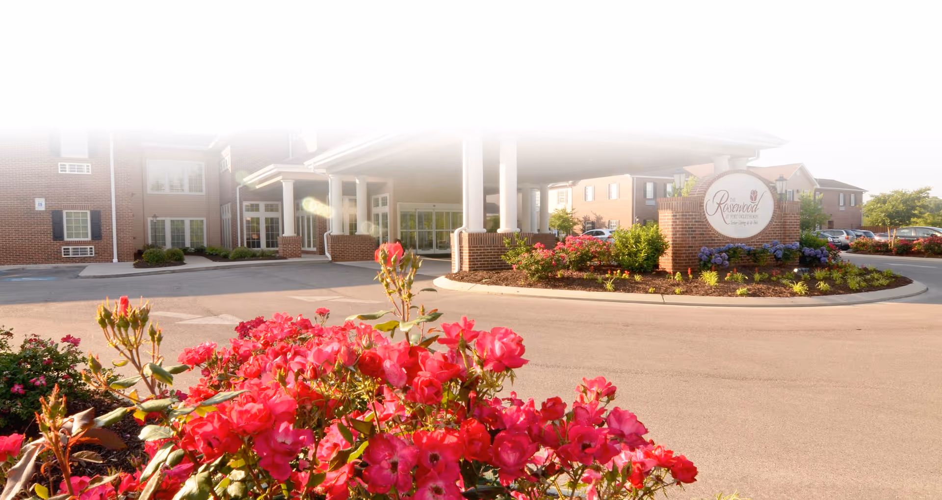 Entrance of The Rosewood at Fort Oglethorpe senior living facility with a covered driveway, brick buildings, and vibrant red and pink flowers in the foreground.