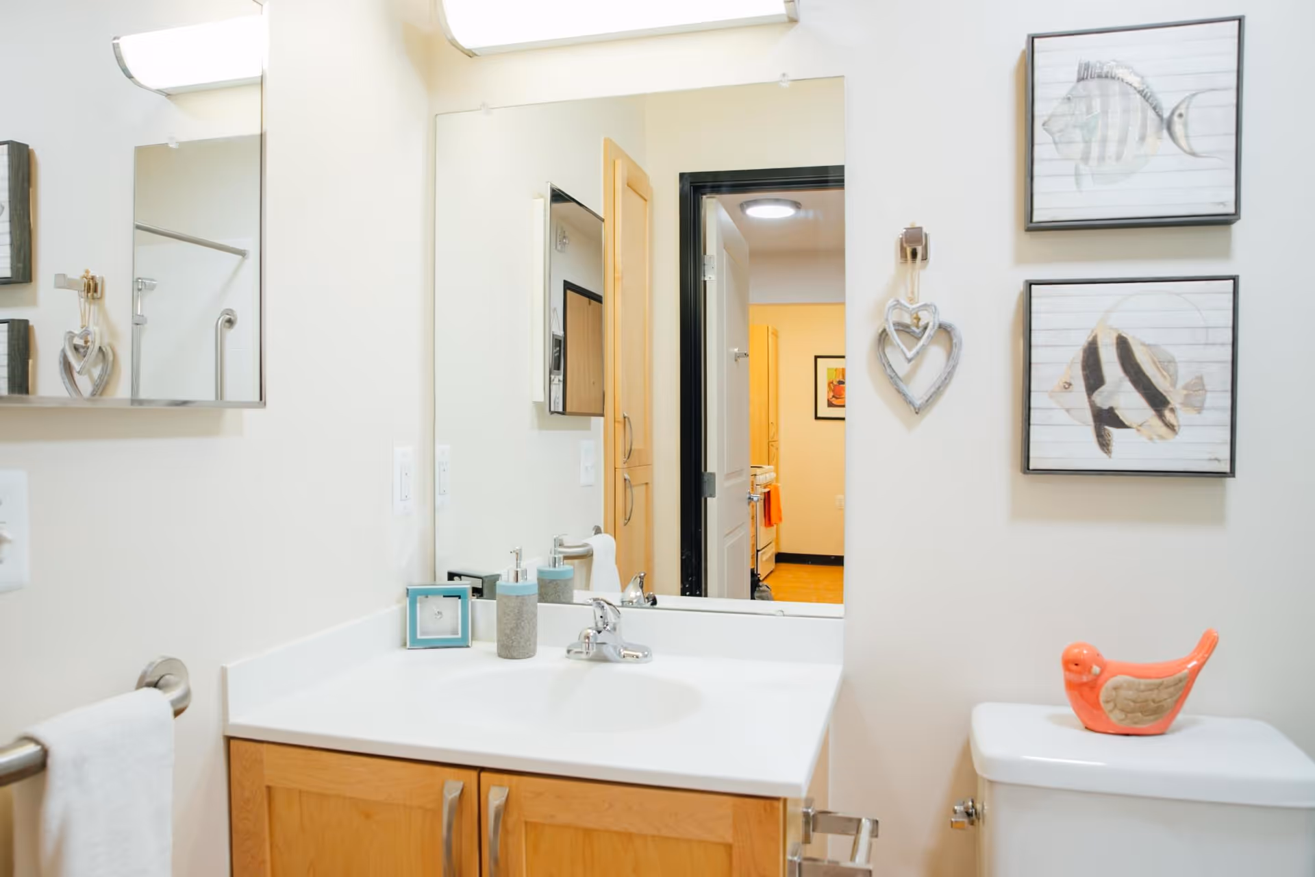 A clean bathroom with a white sink countertop and wooden cabinet below. Above the sink is a large mirror reflecting a hallway and another room. On the countertop are a soap dispenser, a small framed picture, and a toothbrush holder. To the right, there is a white toilet with a decorative orange bird figurine on the tank. The wall above the toilet has two framed fish illustrations and a hanging heart decoration. A towel hangs on a metal bar to the left of the sink.