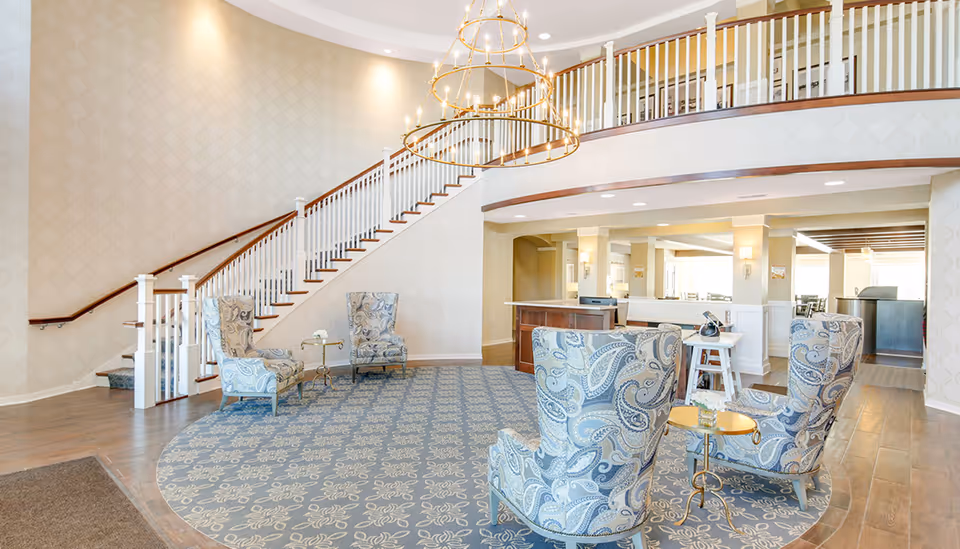 A spacious and elegant senior living facility common area with a large patterned blue rug, four upholstered armchairs with paisley designs, a small round table with a flower vase, a grand staircase with white railings and wooden handrails, and a large chandelier hanging from the ceiling.