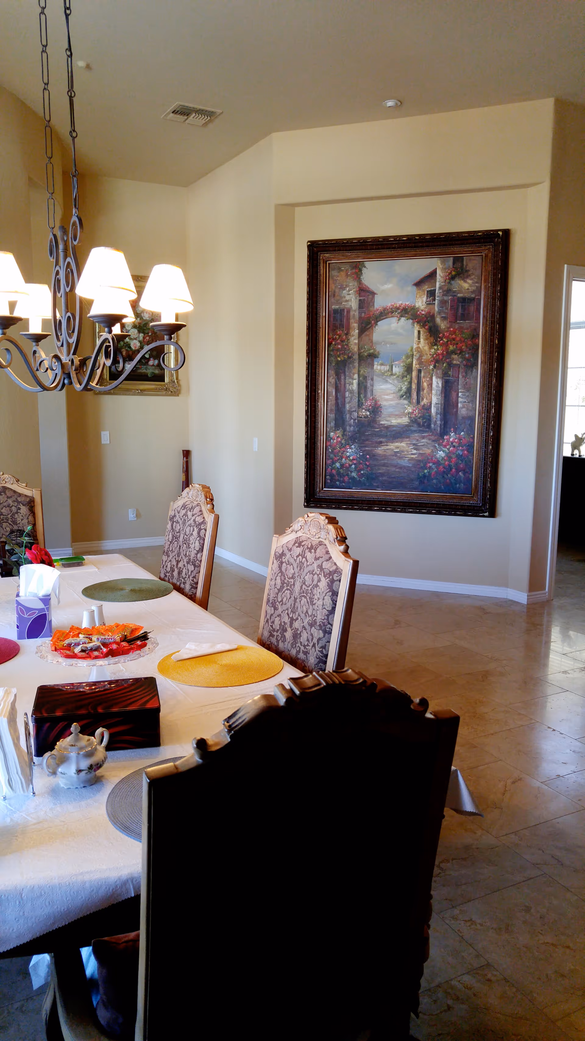 A dining room with a long table covered with a white tablecloth, set with colorful placemats, a tissue box, and a decorative tray. There are ornate chairs around the table and a chandelier with lit lamps hanging above. On the wall, there is a large framed painting depicting a scenic pathway with flowers and buildings.