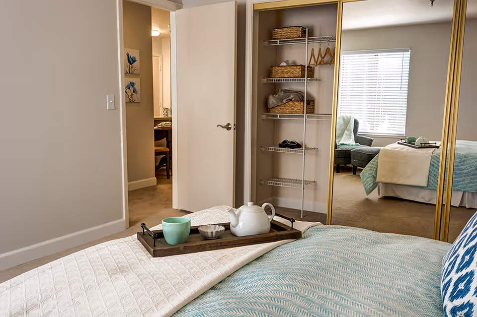 A neatly made bedroom with a tray holding a teapot and cup on the bed, mirrored closet doors, and a windowed sitting area reflected in the mirror.