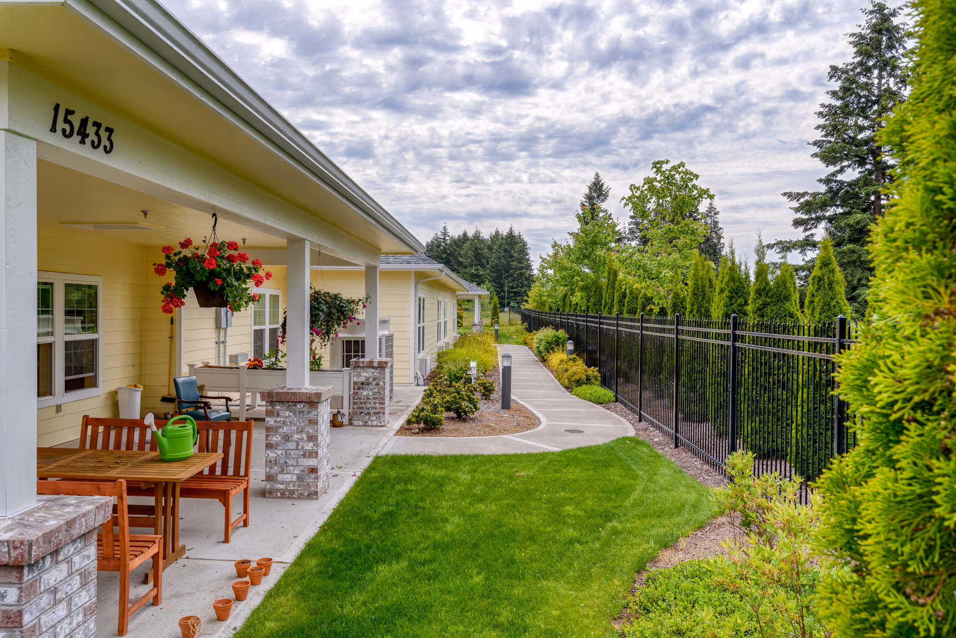 Outdoor view of a senior living facility porch area with a wooden table and chairs, hanging flower pots, and a green watering can on the table. The building is light yellow with white trim, and there is a paved walkway alongside a black metal fence with green trees and shrubs lining the path under a cloudy sky.