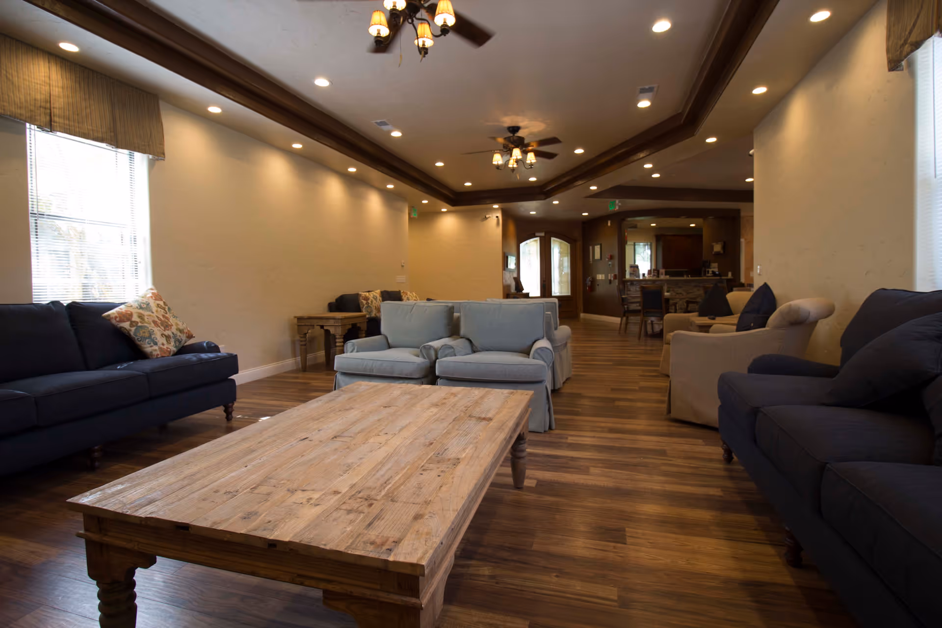 A spacious senior living facility common area with wooden flooring, multiple sofas and armchairs arranged around a large wooden coffee table, soft lighting from ceiling fixtures, and windows with blinds allowing natural light.