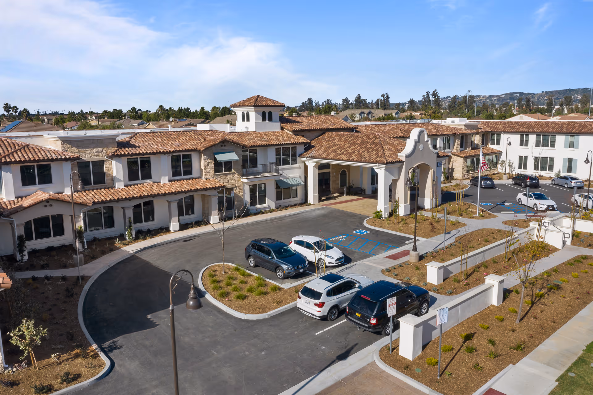 Aerial view of the front entrance and parking area of a Mediterranean-style senior living facility with terracotta tile roofs.