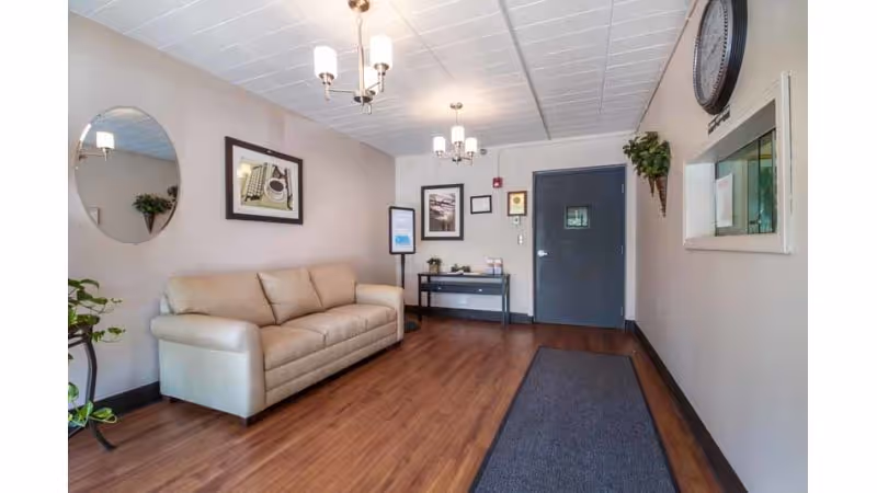 A waiting area with a beige leather sofa against a light-colored wall, a round mirror, framed artwork, a small table with plants and informational materials, a dark door, a wall clock, and a long dark rug on wooden flooring.