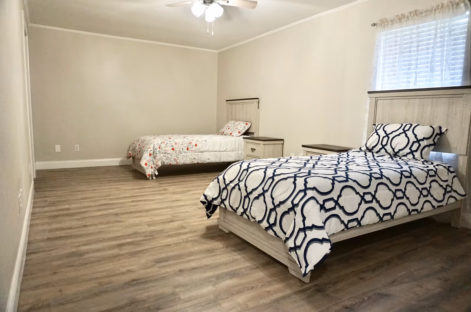 A simple bedroom with two twin beds, each with a different patterned bedspread. The room has light-colored walls, wood flooring, a ceiling fan with lights, and a window with sheer curtains.