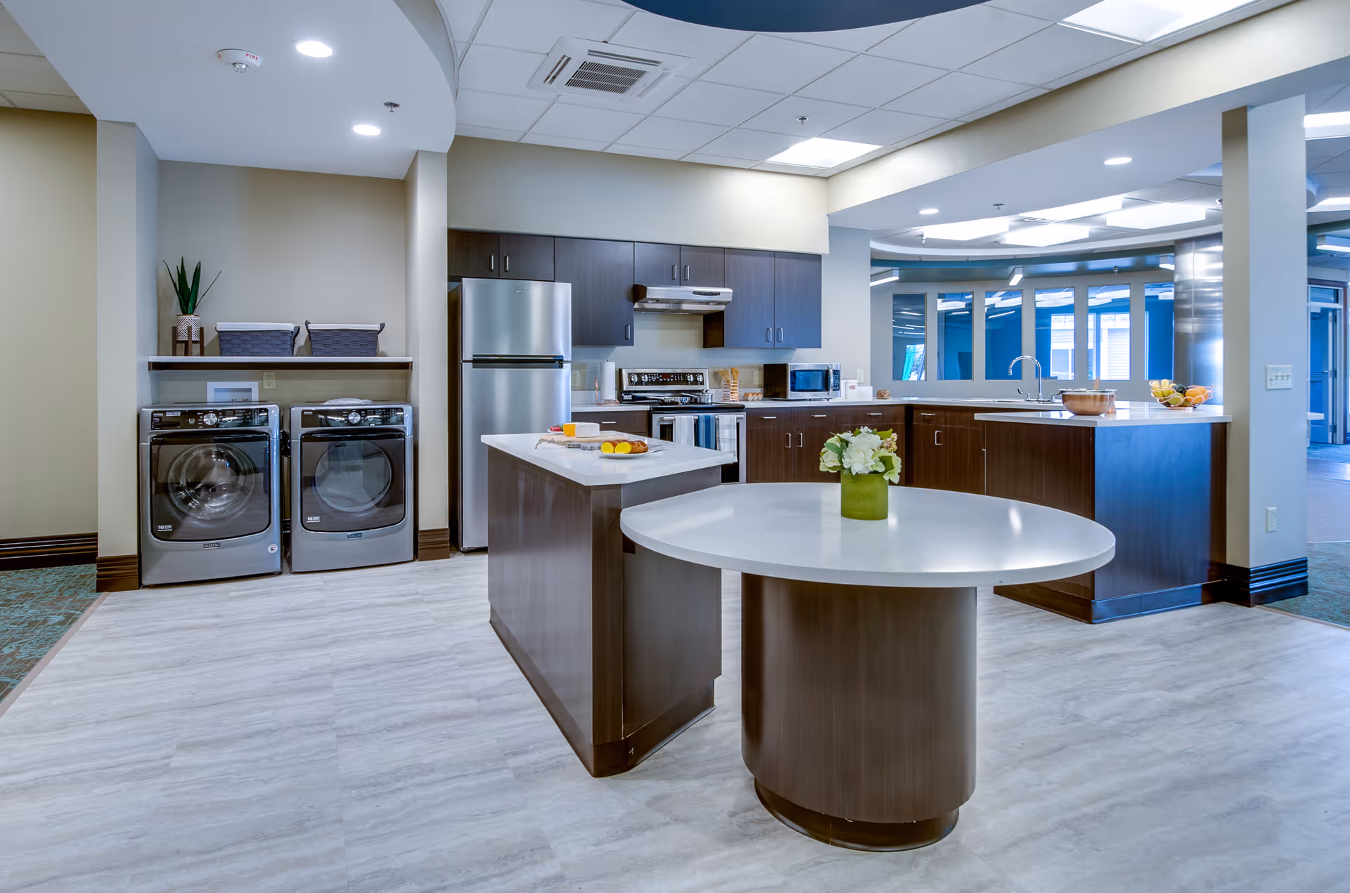 Modern kitchen area with dark wood cabinets, stainless steel refrigerator, stove, microwave, and a double washer and dryer set against a beige wall. The kitchen features two white countertops, one round with a vase of flowers and one rectangular with food items. The ceiling has recessed lighting and an air conditioning vent, and there is a large window opening to another room.