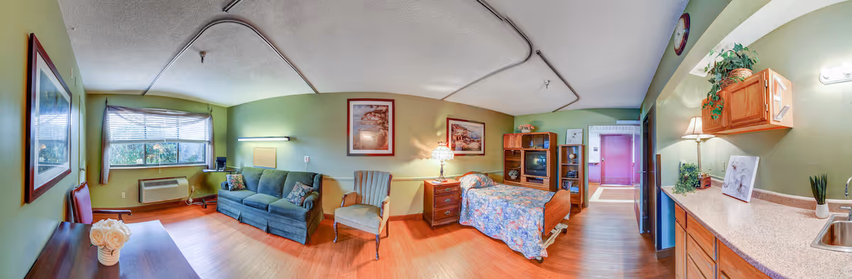 A cozy senior living room and bedroom area with green walls and wooden flooring. The room features a blue sofa, an armchair, a single bed with floral bedding, a wooden nightstand with a lamp, a wooden entertainment center with a TV, and a kitchenette area with a countertop, sink, and cabinets. There are framed pictures on the walls and a window with blinds letting in natural light.