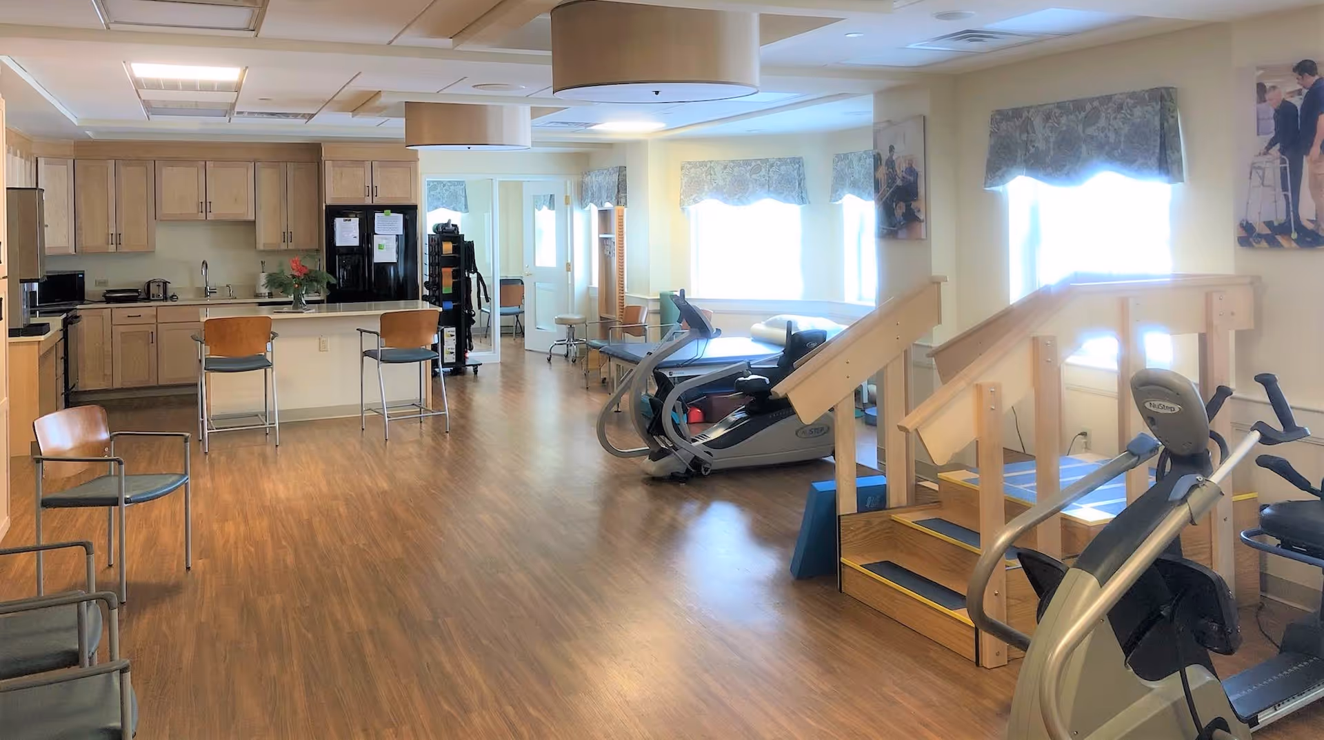 A spacious room with wooden flooring featuring a kitchen area with cabinets, a refrigerator, and a counter with two chairs. The room also contains exercise equipment including a stationary bike, a treadmill, and a wooden staircase for physical therapy. There are several chairs along the left side and windows with patterned valances on the right side, allowing natural light to brighten the space.