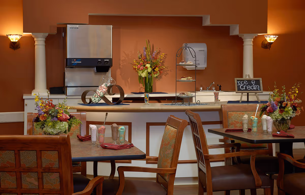 Interior view of a dining area with wooden chairs and tables set with milkshakes and napkins. In the background, there is a counter with an ice dispenser, a tiered tray with desserts, a vase with colorful flowers, and a small chalkboard sign that reads 'Ice Cream'. The walls are painted a warm brown color with decorative columns and wall sconces providing lighting.