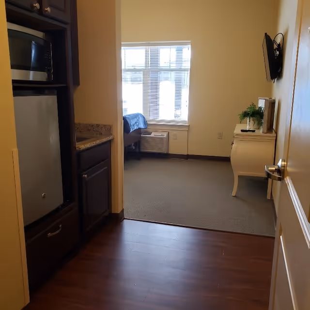 View from a small kitchen area with a microwave and mini refrigerator on the left, looking into a bedroom with a bed near a window with blinds. A white dresser with a plant and a mounted TV are visible on the right side of the bedroom.