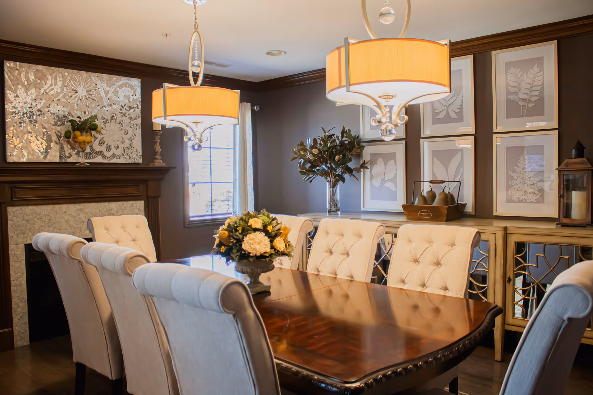 A formal dining room with a polished wooden table surrounded by eight beige upholstered chairs. Two large pendant lights hang above the table. A floral centerpiece is placed in the middle of the table. The room features a window with curtains, a decorative fireplace with a large ornate mirror above it, and a sideboard with framed botanical prints and decorative items.
