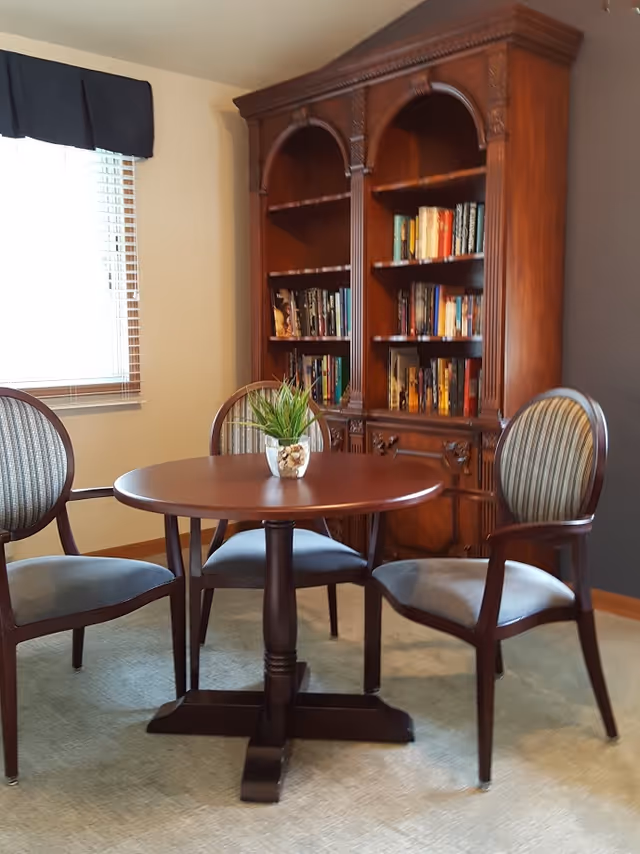 A cozy interior room featuring a round wooden table with a small potted plant centerpiece, surrounded by three upholstered wooden chairs. Behind the table is a large wooden bookshelf filled with books. A window with blinds and a dark valance is on the left wall.