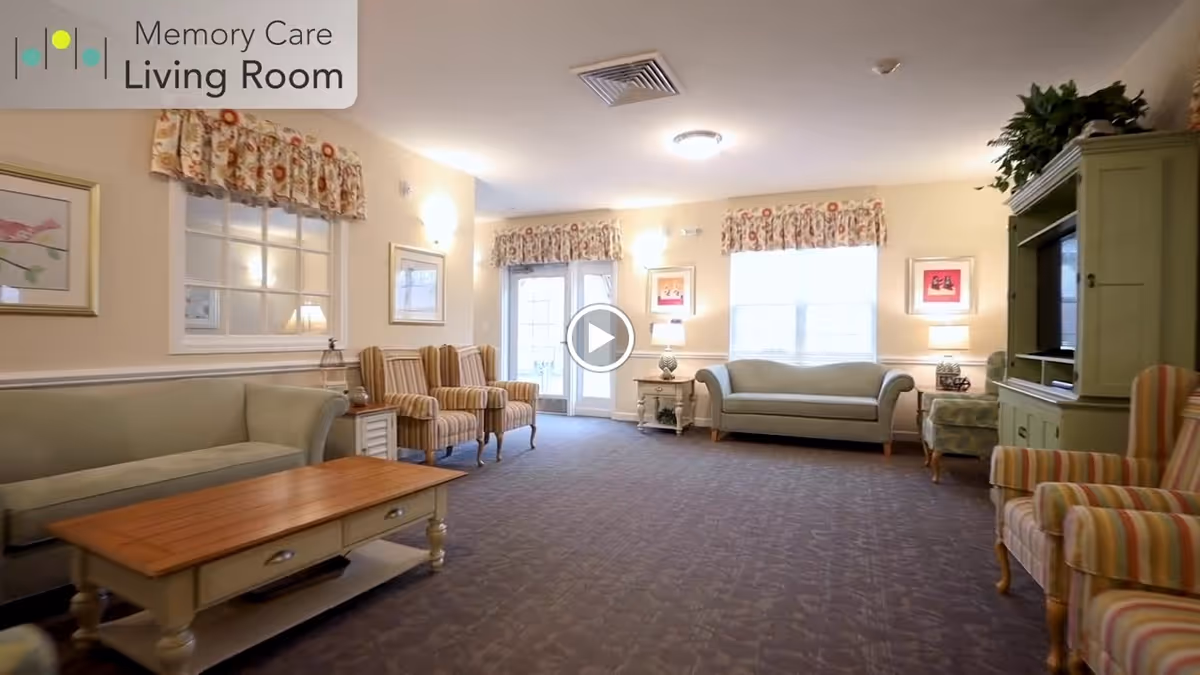 Spacious, softly lit memory care living room with sofas, striped armchairs, a coffee table, and a TV cabinet.