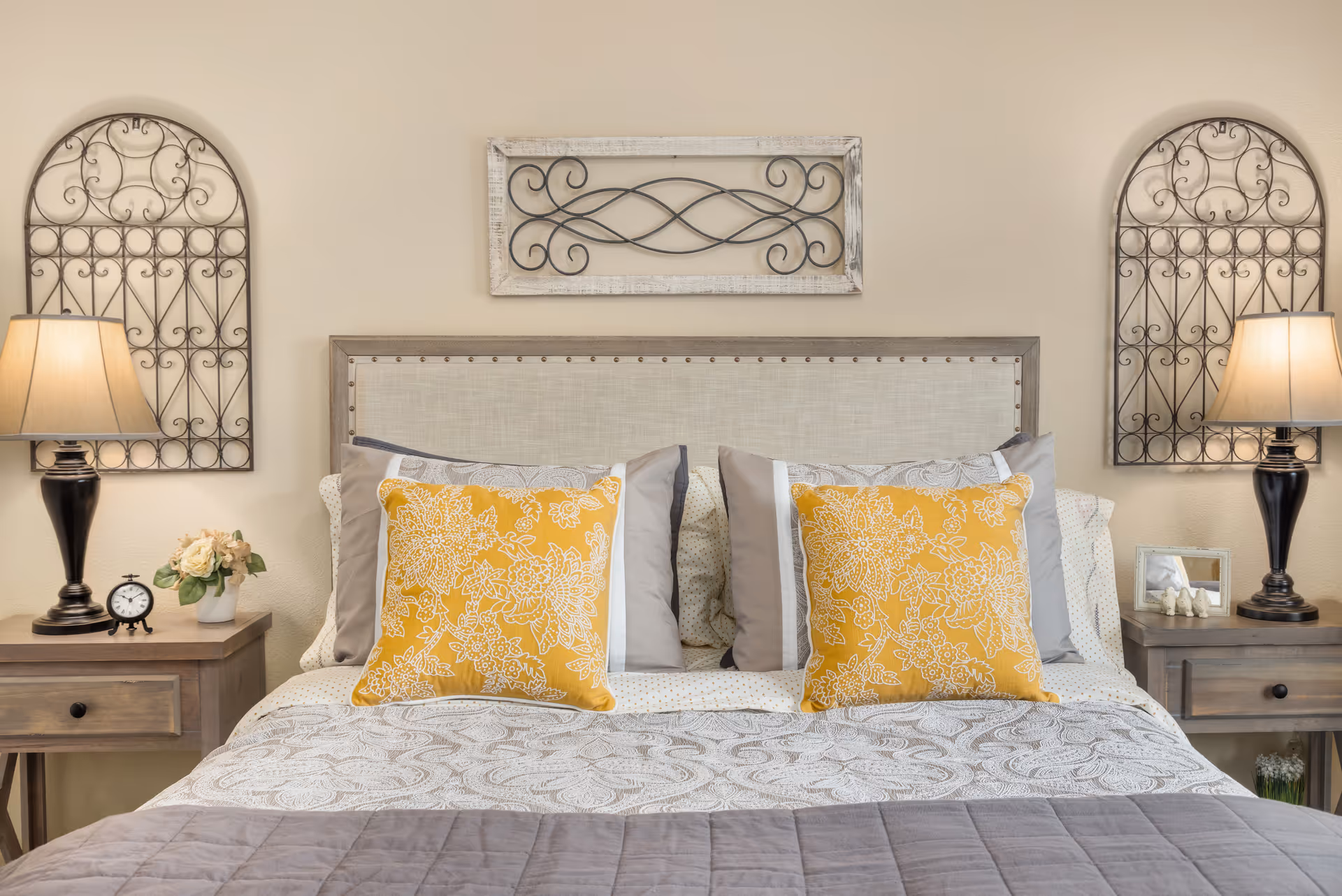 A neatly made bed with a beige upholstered headboard, decorated with two yellow floral pillows and multiple gray and white pillows. On each side of the bed are wooden nightstands with black lamps, a small clock, a flower vase, and a picture frame. Above the headboard are three decorative wrought iron wall hangings.