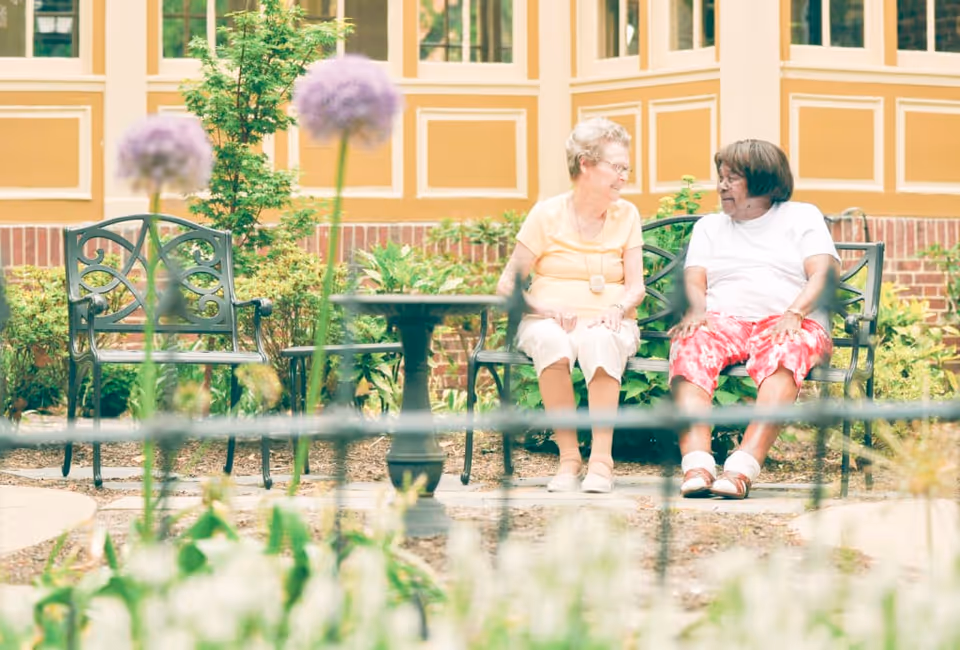 Two elderly women sitting and talking on a metal bench in a garden area with flowers and greenery, in front of a building with large windows and yellow walls.