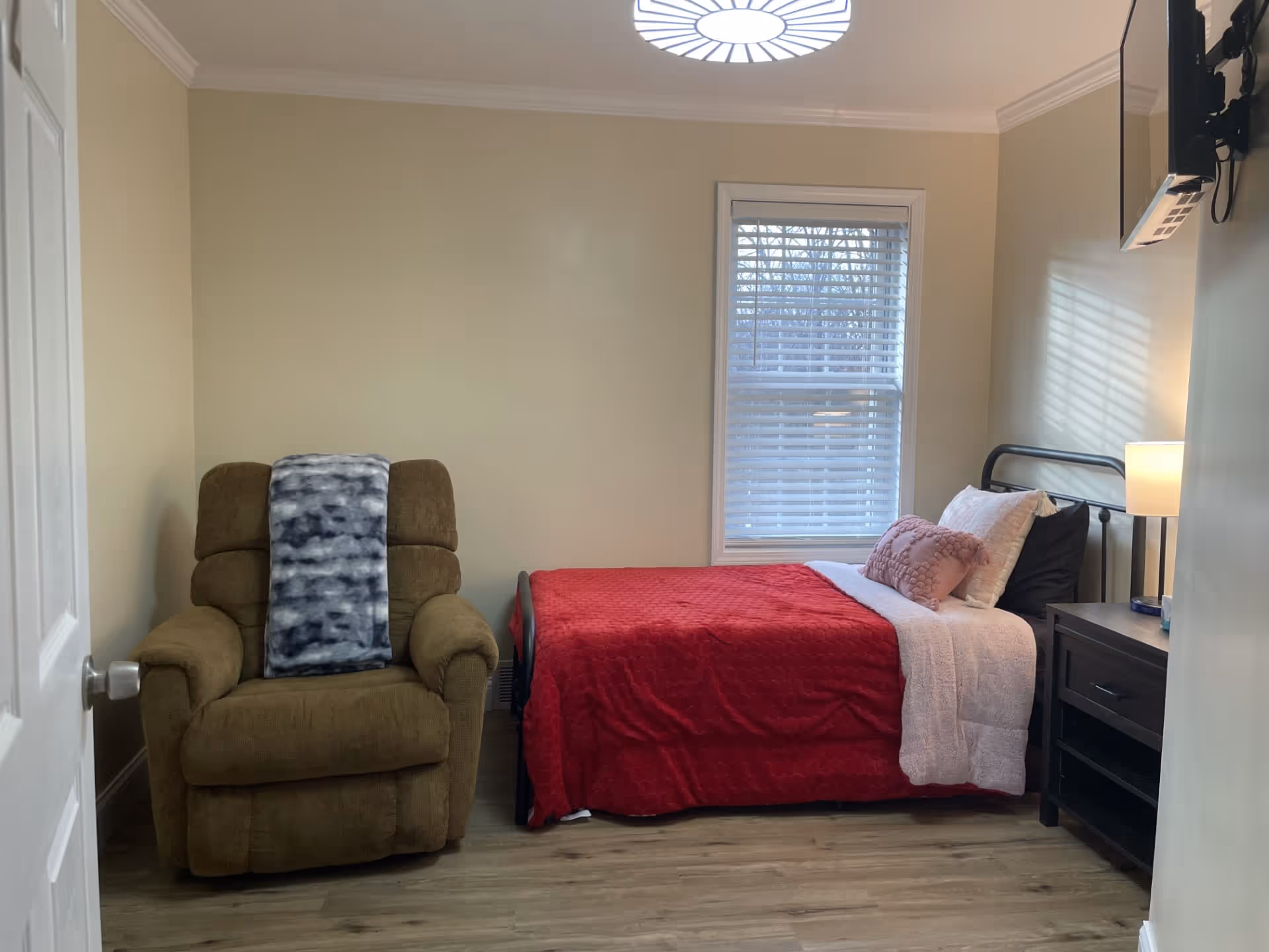 A small bedroom with a single bed covered in a red blanket and pink pillows, a brown recliner chair with a gray and white blanket draped over it, a window with white blinds, a dark wooden nightstand with a lamp, and a wall-mounted TV.