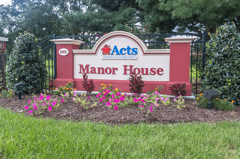 A landscaped entrance sign for Manor House, part of Acts Retirement Life Communities, surrounded by green grass, colorful flowers, and bushes with trees in the background.