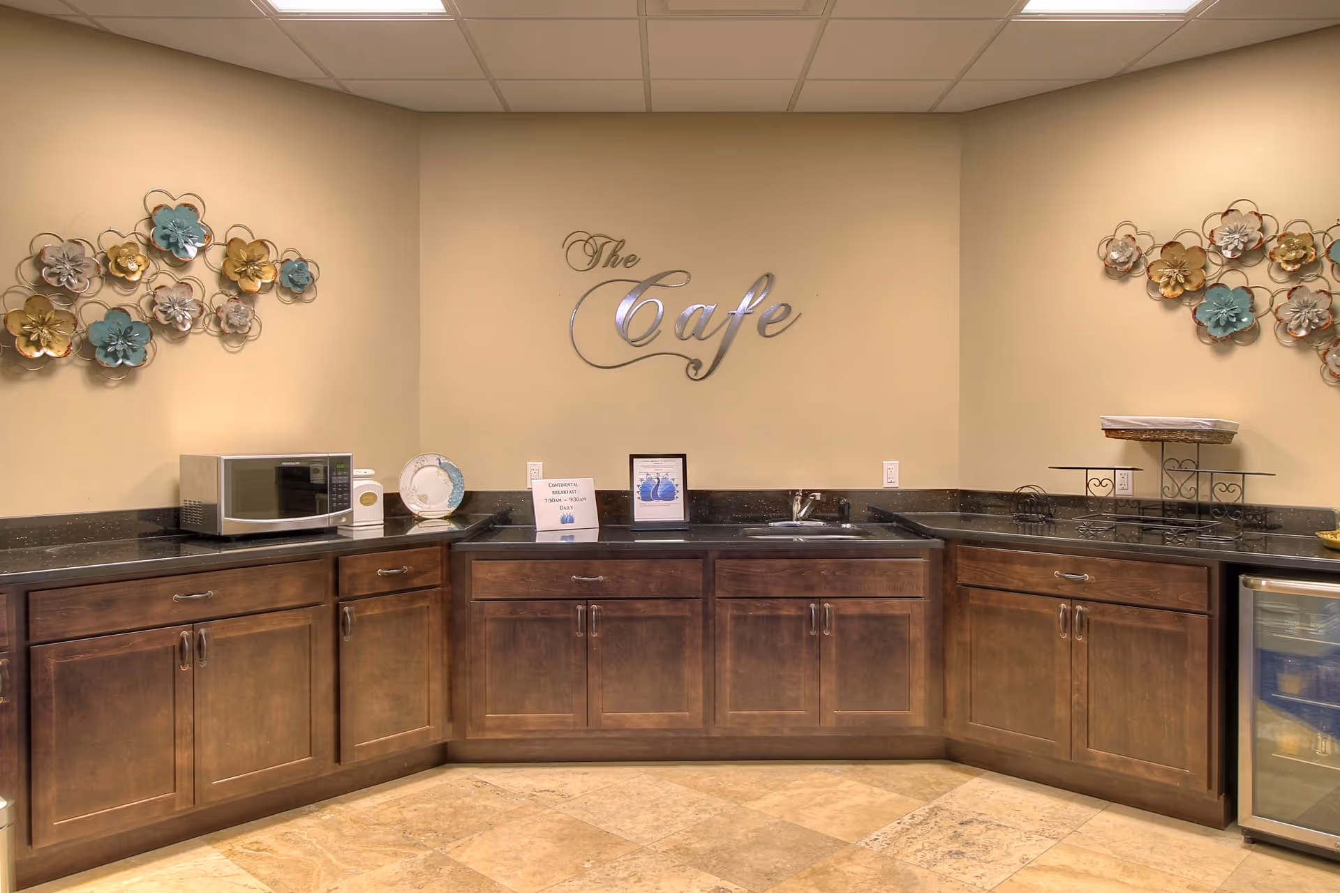 A small cafe-style kitchenette with dark wood cabinets, a sink and microwave beneath a wall sign reading "The Cafe" and floral wall decorations.