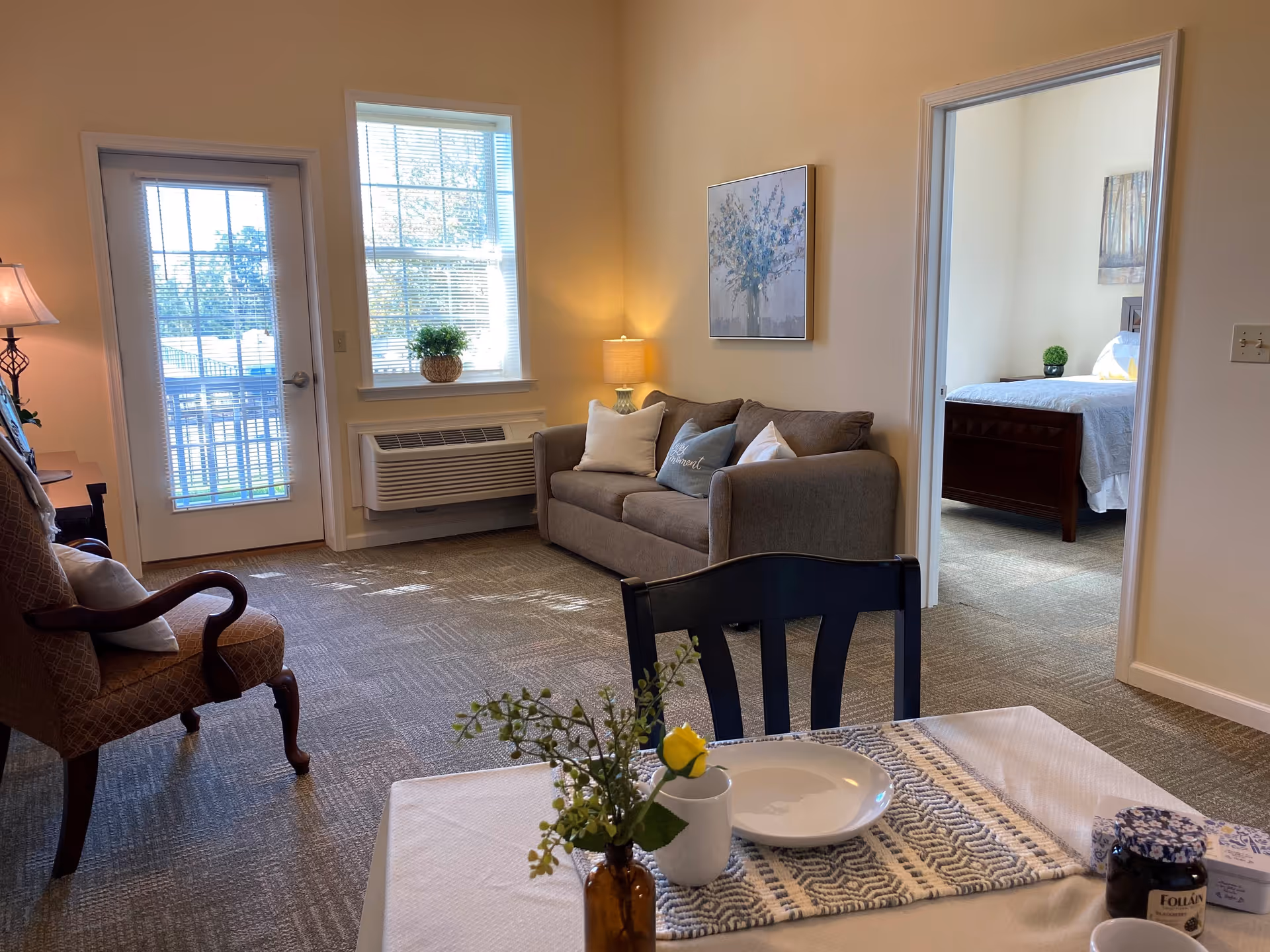 A cozy living area in a senior living facility featuring a gray sofa with decorative pillows, a wooden armchair with a cushion, a small table set with a plate, cup, and a vase with flowers. There is a window with blinds and a door leading outside. In the background, a bedroom with a bed and nightstand is visible through an open doorway.