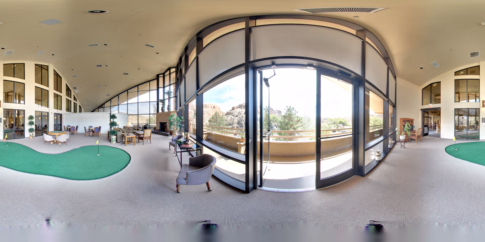 Spacious senior living common area with an indoor putting green, seating groups and large floor-to-ceiling windows opening to a balcony with rocky outdoor views.