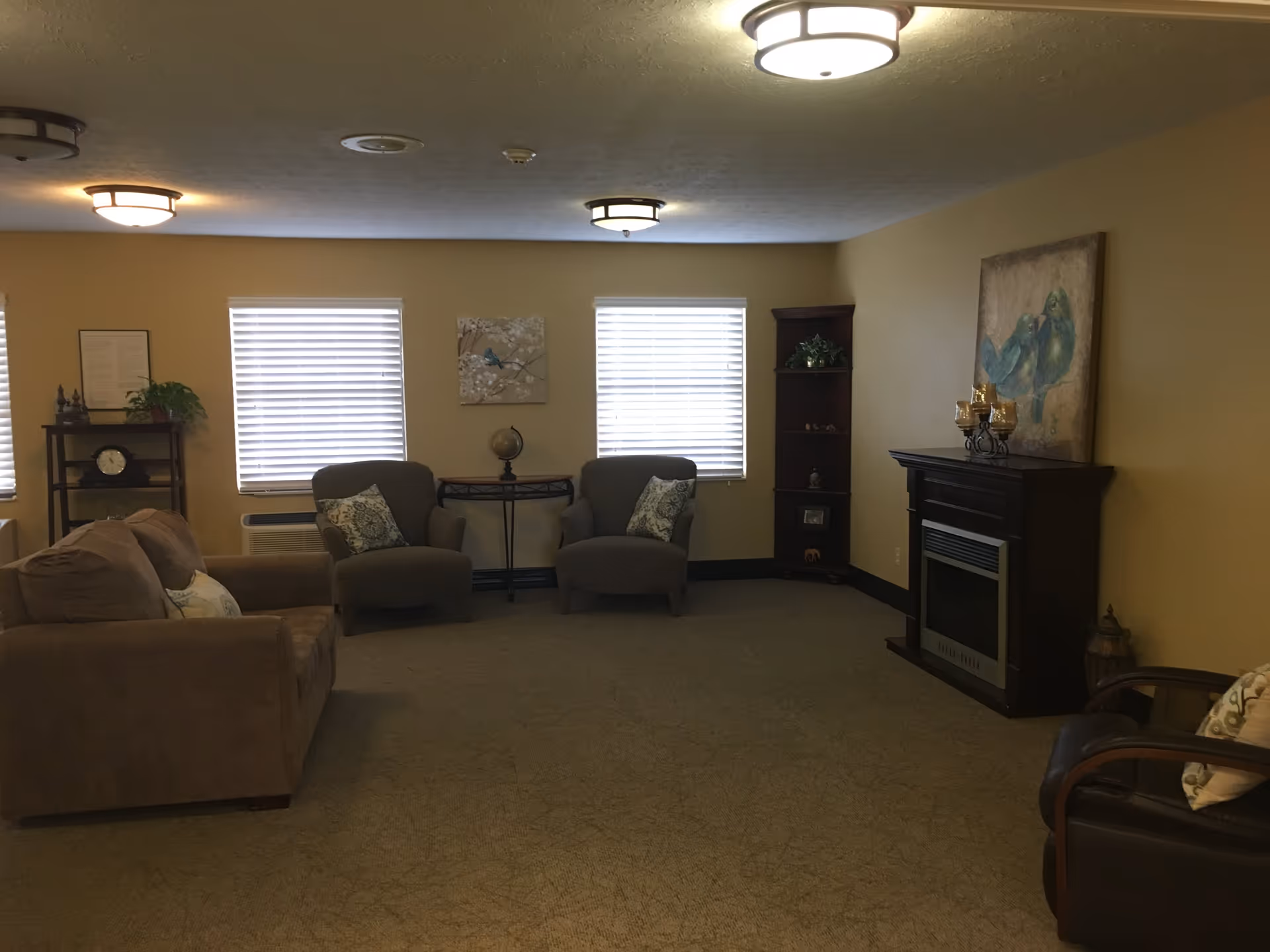A cozy living room with beige walls and carpet, featuring a brown sofa, two gray armchairs with patterned pillows, a dark wooden corner shelf with decorative items, a small table with a globe between the armchairs, and a dark wooden electric fireplace with a painting of blue birds above it. The room is lit by ceiling lights and has three windows with white blinds.