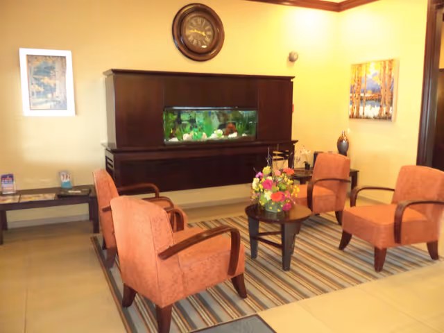 A cozy sitting area with four orange upholstered armchairs arranged around a small round table with a colorful flower arrangement. Behind the chairs is a wall-mounted dark wooden cabinet with a built-in aquarium. A round clock hangs above the cabinet, and two framed paintings are on the walls. The floor has a striped area rug and beige tiles.
