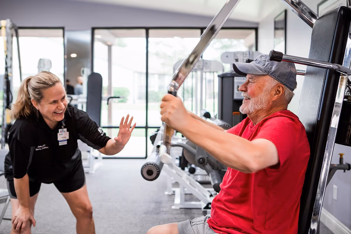 An elderly man in a red shirt and gray cap is using a weight machine in a gym while a female fitness trainer in black sportswear encourages him with a smile and raised hand. The gym has large windows letting in natural light and various exercise equipment in the background.