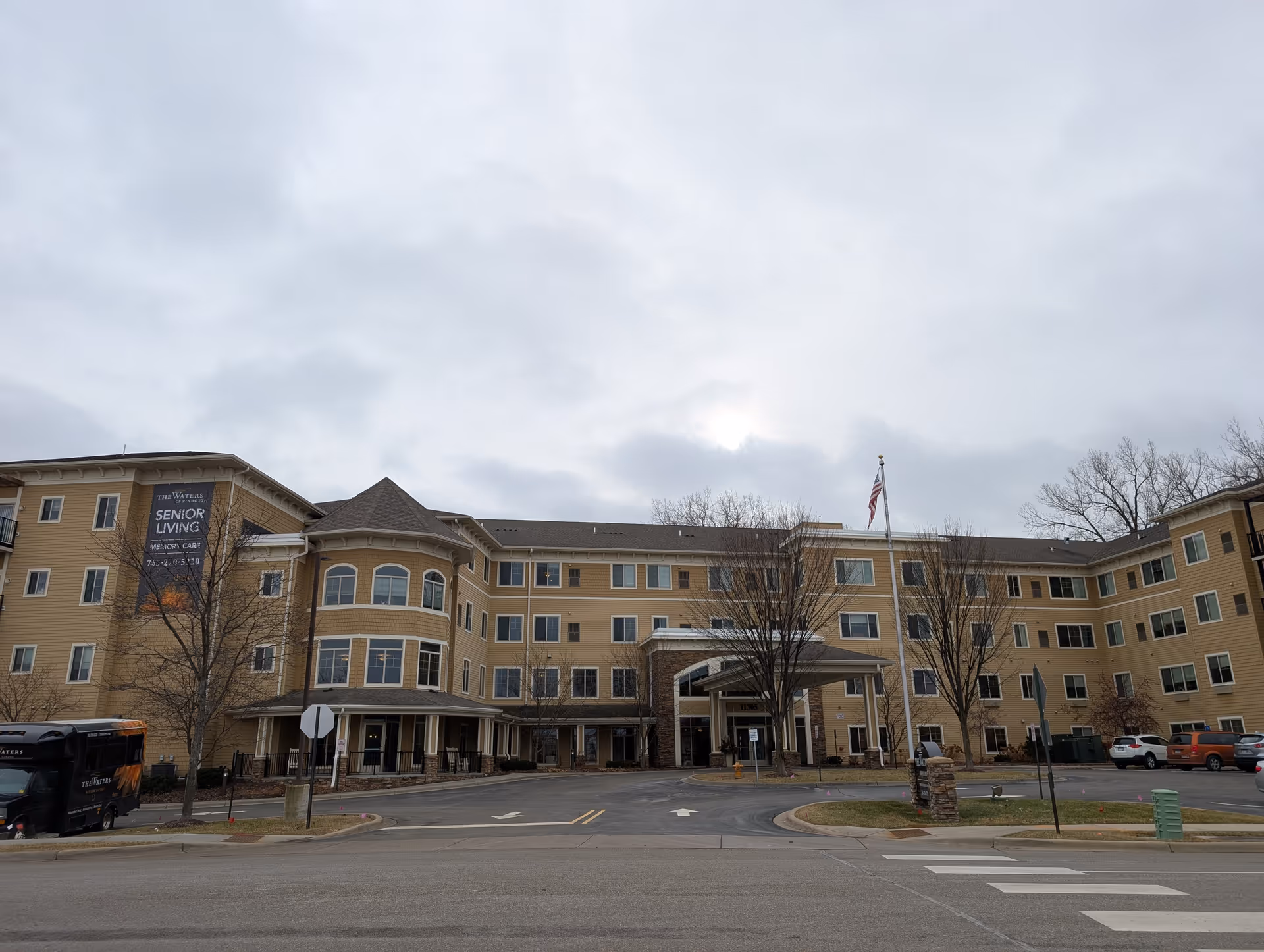 Four-story beige senior living facility with a covered main entrance and an American flag in front under a cloudy sky.
