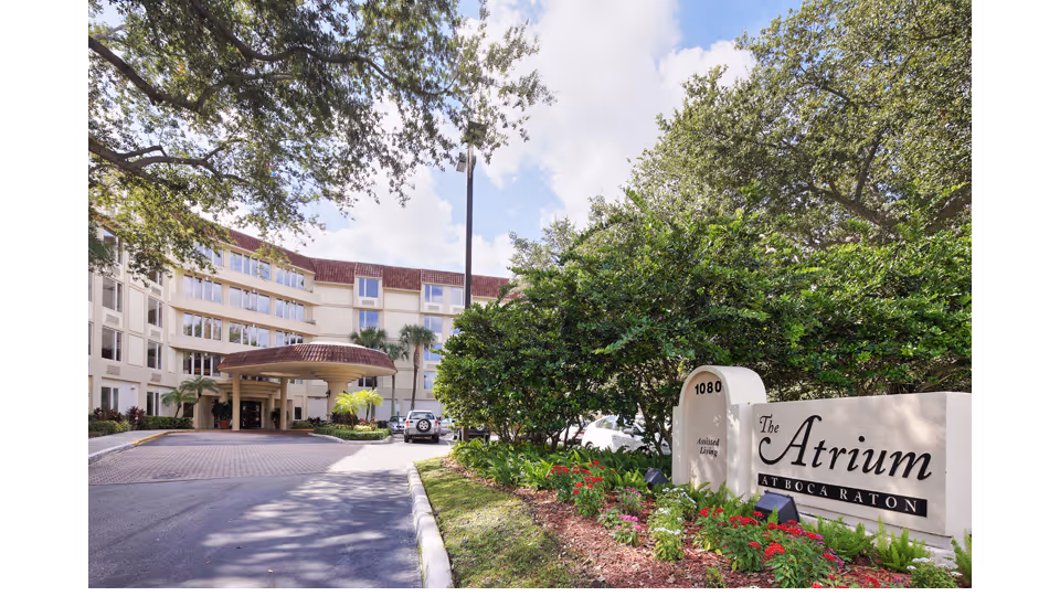 Exterior view of The Atrium at Boca Raton senior living facility showing the building entrance with a covered driveway, surrounded by trees and landscaped greenery under a partly cloudy sky.