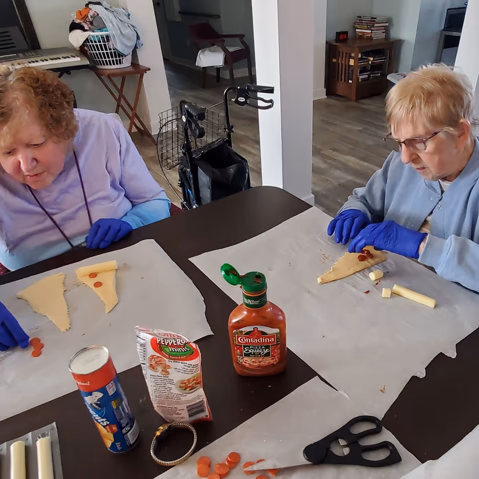Two elderly women wearing blue gloves are seated at a table preparing small pizza slices with dough, mini pepperoni, cheese sticks, and pizza sauce in a room with wooden flooring and furniture.