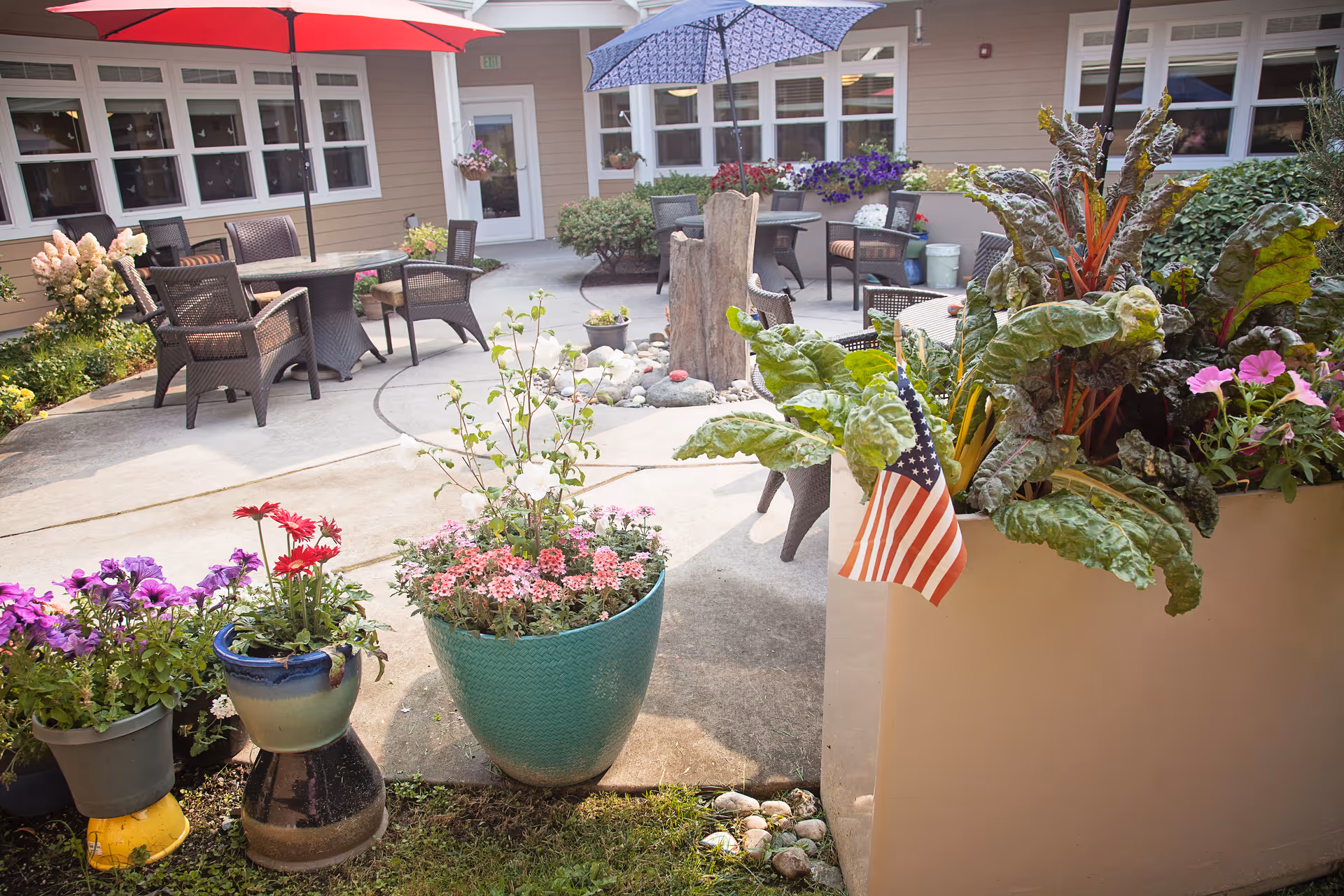 Outdoor courtyard area with several potted plants and flowers, including a large planter with leafy greens and a small American flag. There are multiple tables with chairs and umbrellas providing shade. The courtyard is surrounded by a building with windows and a door.