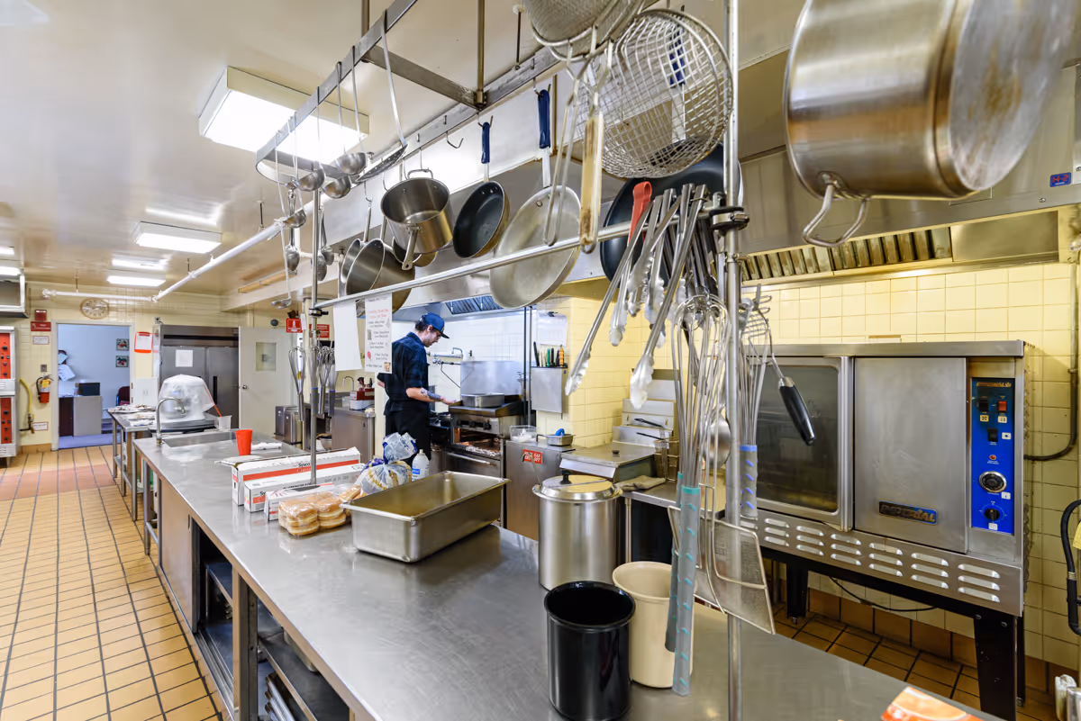 A commercial kitchen with stainless steel counters and hanging pots, pans, and utensils. A person wearing a cap and apron is cooking at a stove. The kitchen has tiled floors and walls, with various kitchen equipment including ovens and storage racks.