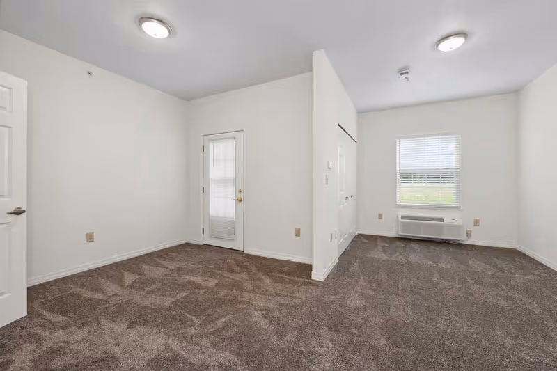 Empty room with beige carpet and white walls, featuring a window with blinds, a door with a glass panel, and a small closet with double doors. The room is well-lit with ceiling lights and has a wall-mounted air conditioning unit below the window.