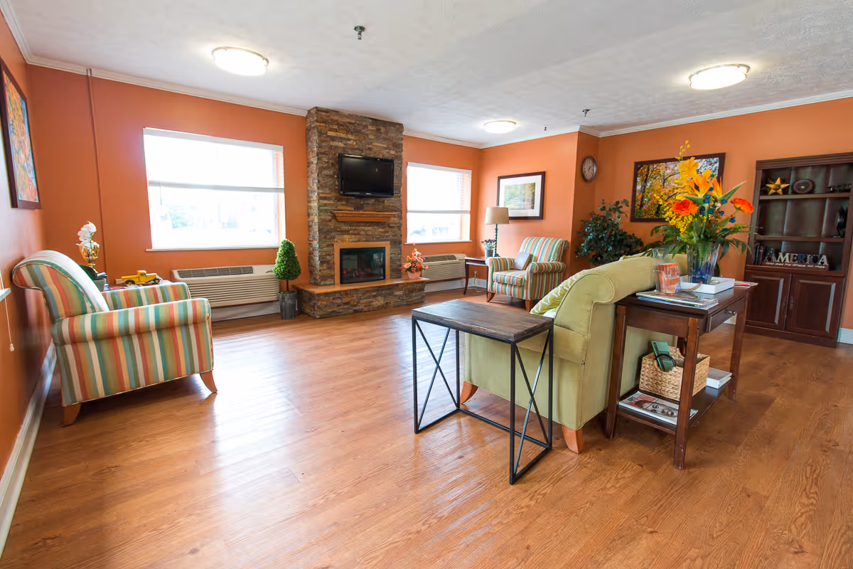 Bright communal living room with striped armchairs, a green sofa, coffee tables, a stone fireplace with mounted TV, and floral arrangements.