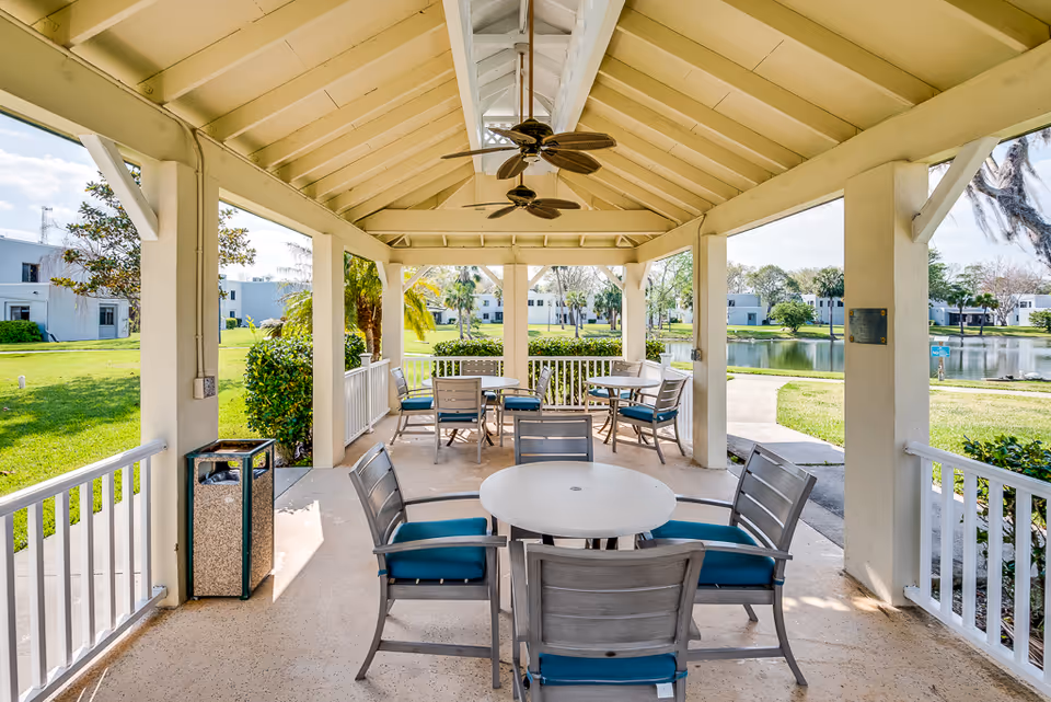 Covered outdoor seating area with round tables and chairs featuring blue cushions, ceiling fans, and a view of a pond and surrounding greenery in a senior living community.