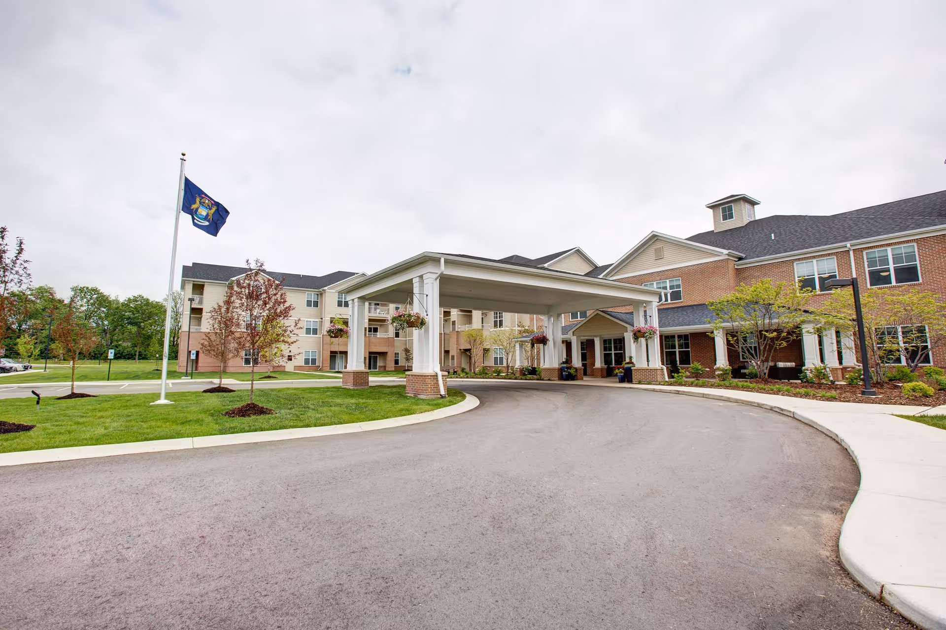 Exterior view of StoryPoint Portage senior living facility showing a large covered entrance with white pillars, a curved driveway, landscaped green lawn with small trees, and a flagpole with a flag. The building has multiple windows and a combination of brick and siding exterior under a cloudy sky.