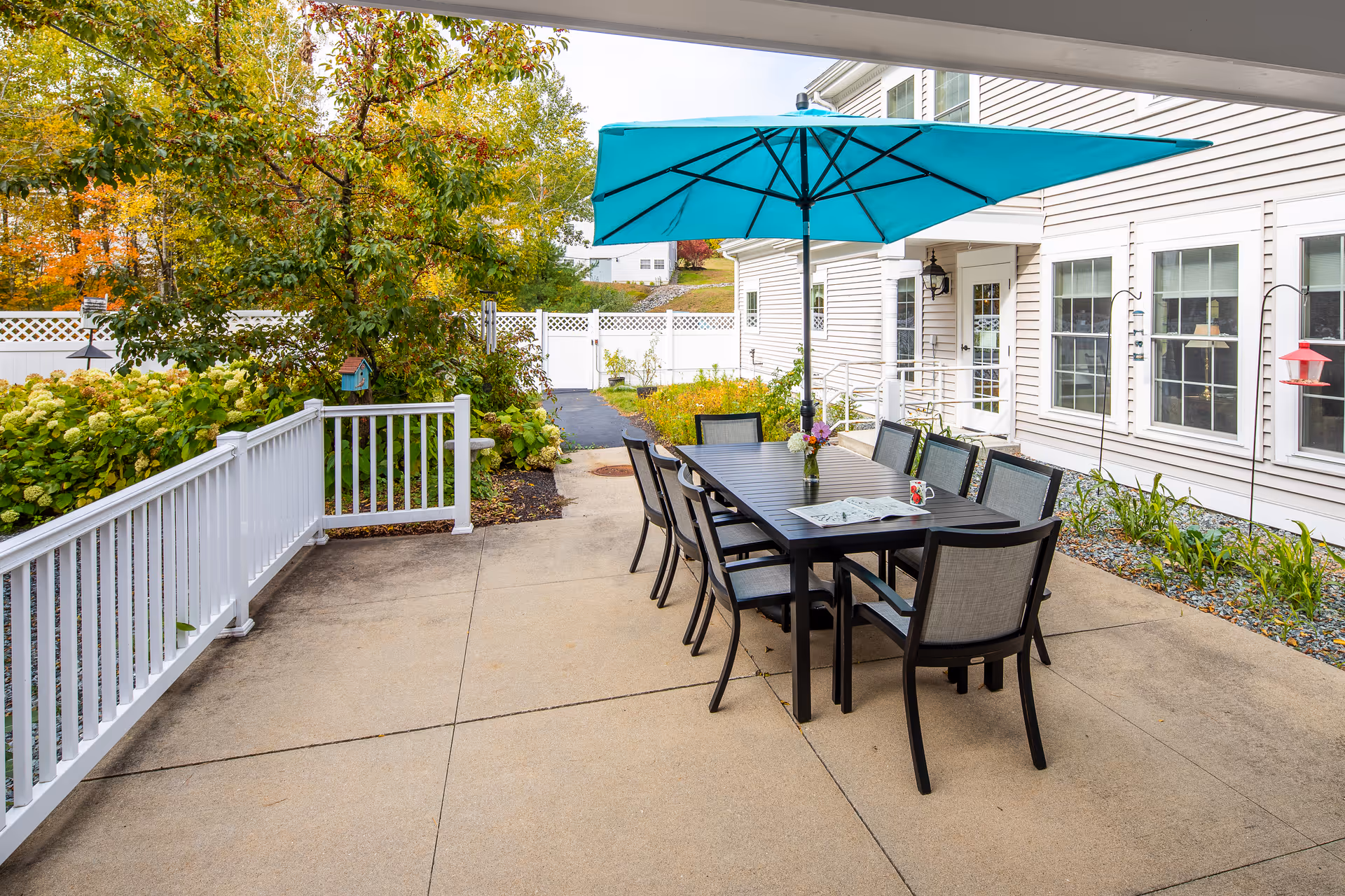 Outdoor patio area with a long black table surrounded by eight chairs. A large blue umbrella shades the table. There is a vase with flowers and a newspaper on the table. The patio is adjacent to a light-colored building with multiple windows and a door. Surrounding the patio are plants, bushes, and a white fence, with trees showing autumn foliage in the background.