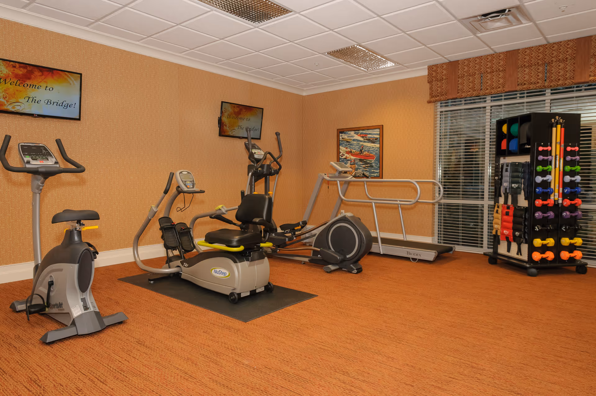 A fitness room with exercise equipment including a stationary bike, a recumbent stepper, an elliptical machine, a treadmill, and a rack of colorful dumbbells and exercise bands. The walls are beige with two mounted TVs displaying a welcome message for The Bridge. There is a window with blinds and a patterned valance.