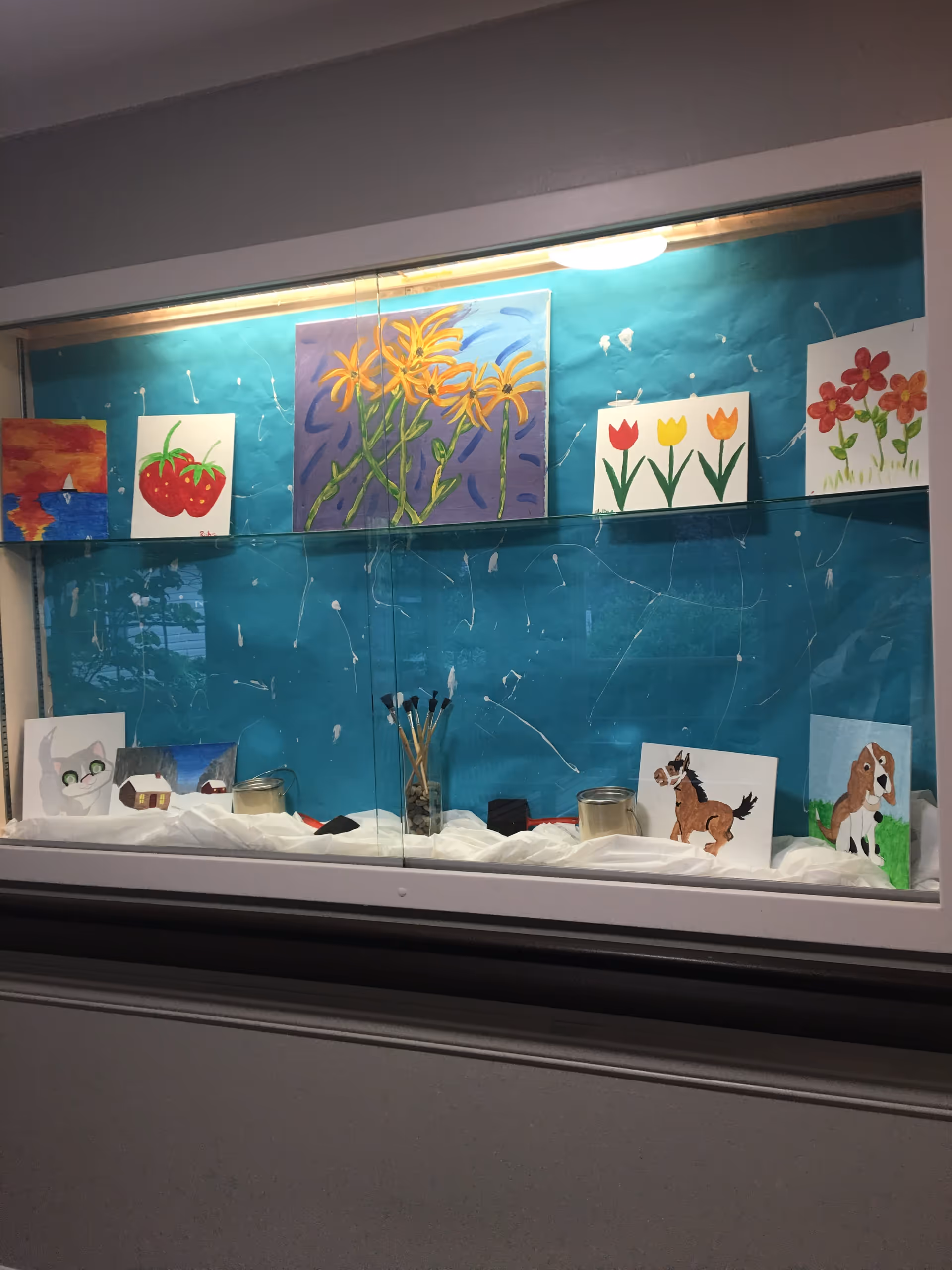 A display case with various colorful paintings including flowers, strawberries, a cat, a horse, and a dog. Paintbrushes are placed in a glass container in the center of the display, which is set against a blue background.