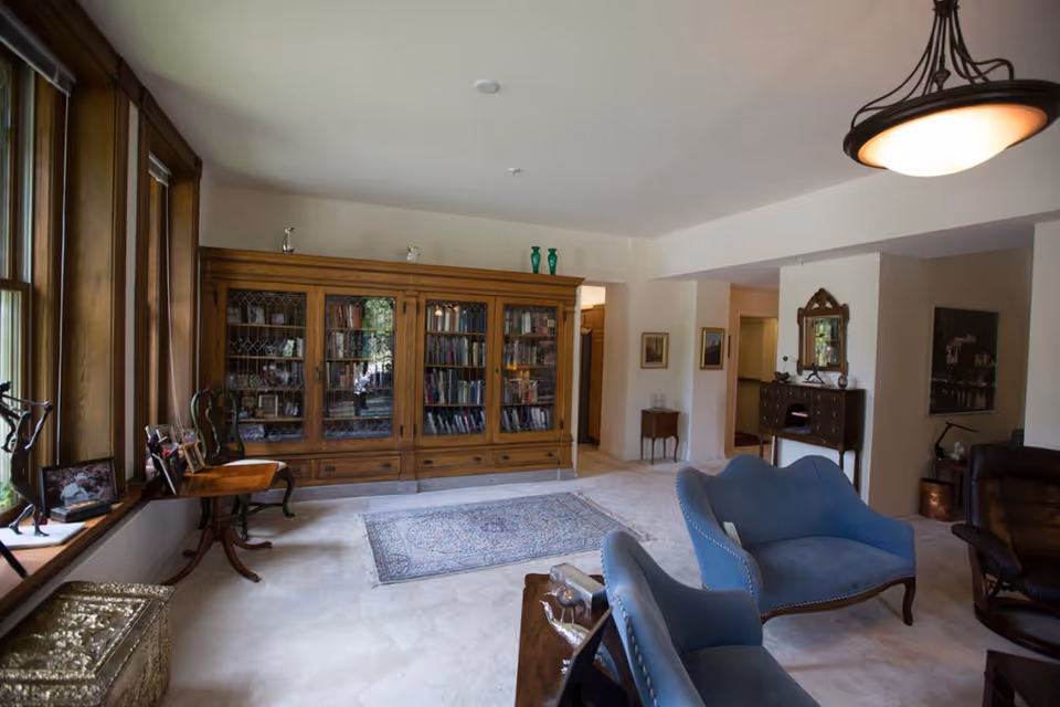 A spacious living room with large wooden-framed windows on the left, a wooden cabinet filled with books and decorative items against the far wall, two blue upholstered chairs in the center, a small wooden table with framed photos near the windows, a patterned rug on the carpeted floor, and additional furniture including a wooden sideboard with a mirror and a leather chair on the right.