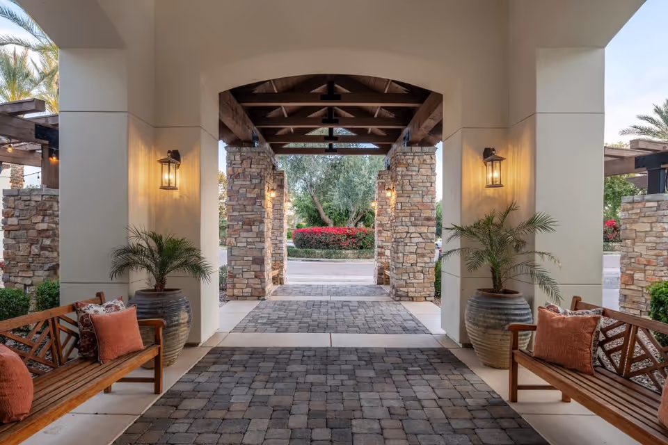 Covered outdoor walkway with stone pillars and wooden benches on either side, each bench adorned with orange and patterned cushions. Large potted plants are placed next to the benches, and warm wall-mounted lanterns illuminate the area. In the background, there are trees and bushes with red flowers.