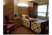A private nursing facility bedroom with a single bed covered by a patchwork quilt, a bedside table with flowers, an armchair, and a window.