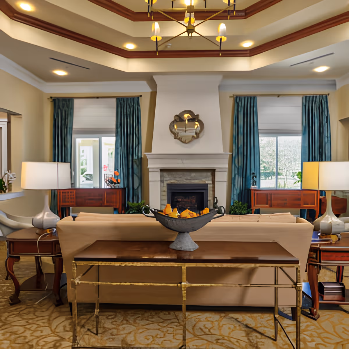 A cozy living room with a beige sofa facing a white fireplace. The room features blue curtains on two windows, a decorative mirror above the fireplace, two side tables with lamps, and a wooden console table behind the sofa with a decorative bowl. The ceiling has recessed lighting and a chandelier.