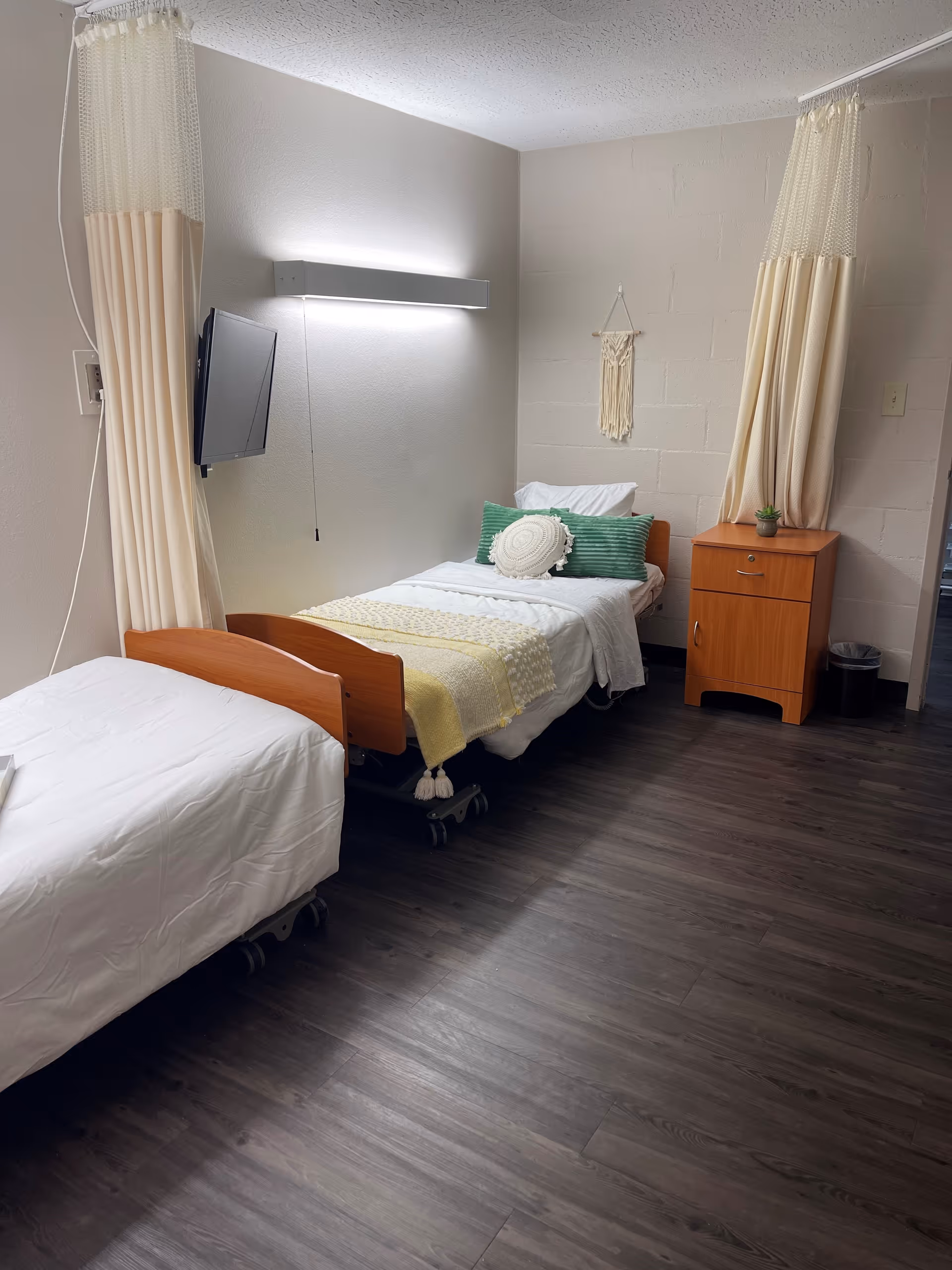 A nursing facility room with two hospital beds, one made with white bedding and the other with white and yellow bedding, green and white pillows. There is a wooden nightstand with a small plant on it, a wall-mounted TV, a wall light, and privacy curtains hanging from the ceiling.