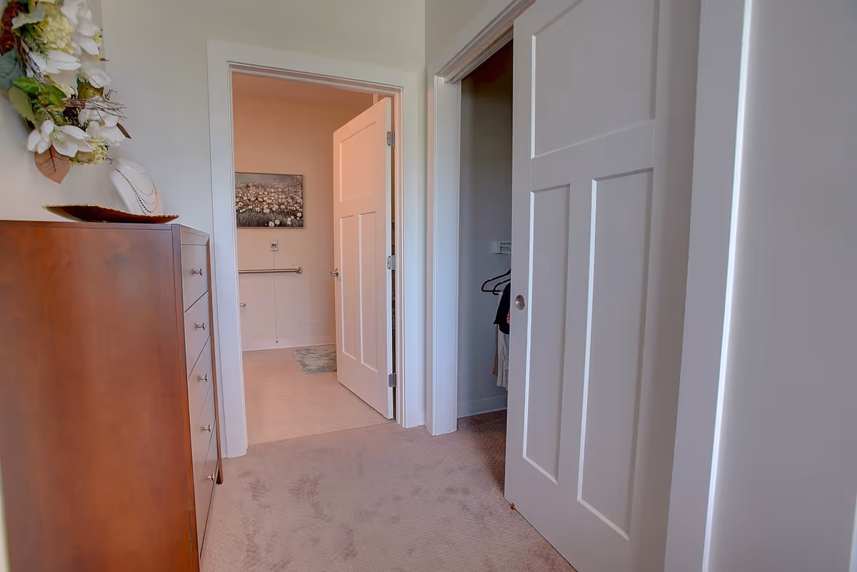 View of a hallway in a senior living facility showing a wooden dresser with decorative items on top, an open door leading to a bathroom with a floral painting on the wall, and a partially open closet door with clothes hanging inside.