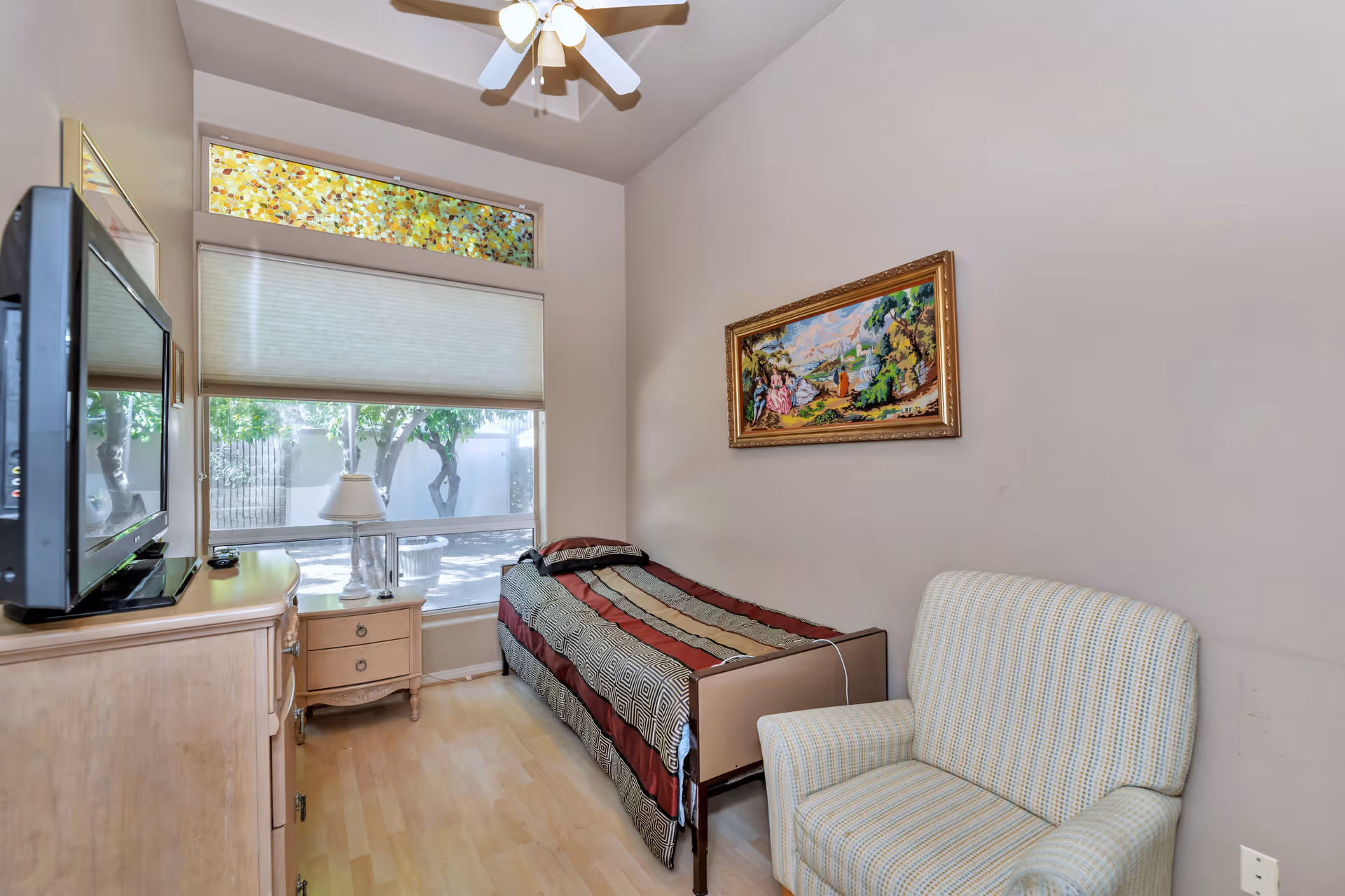 A small bedroom with a single bed covered with a patterned blanket, a cushioned armchair, a wooden dresser with a flat-screen TV on top, and a nightstand with a lamp. The room has a large window with a stained glass panel above it, letting in natural light. A ceiling fan with lights is mounted on the ceiling, and a framed painting hangs on the wall above the bed.