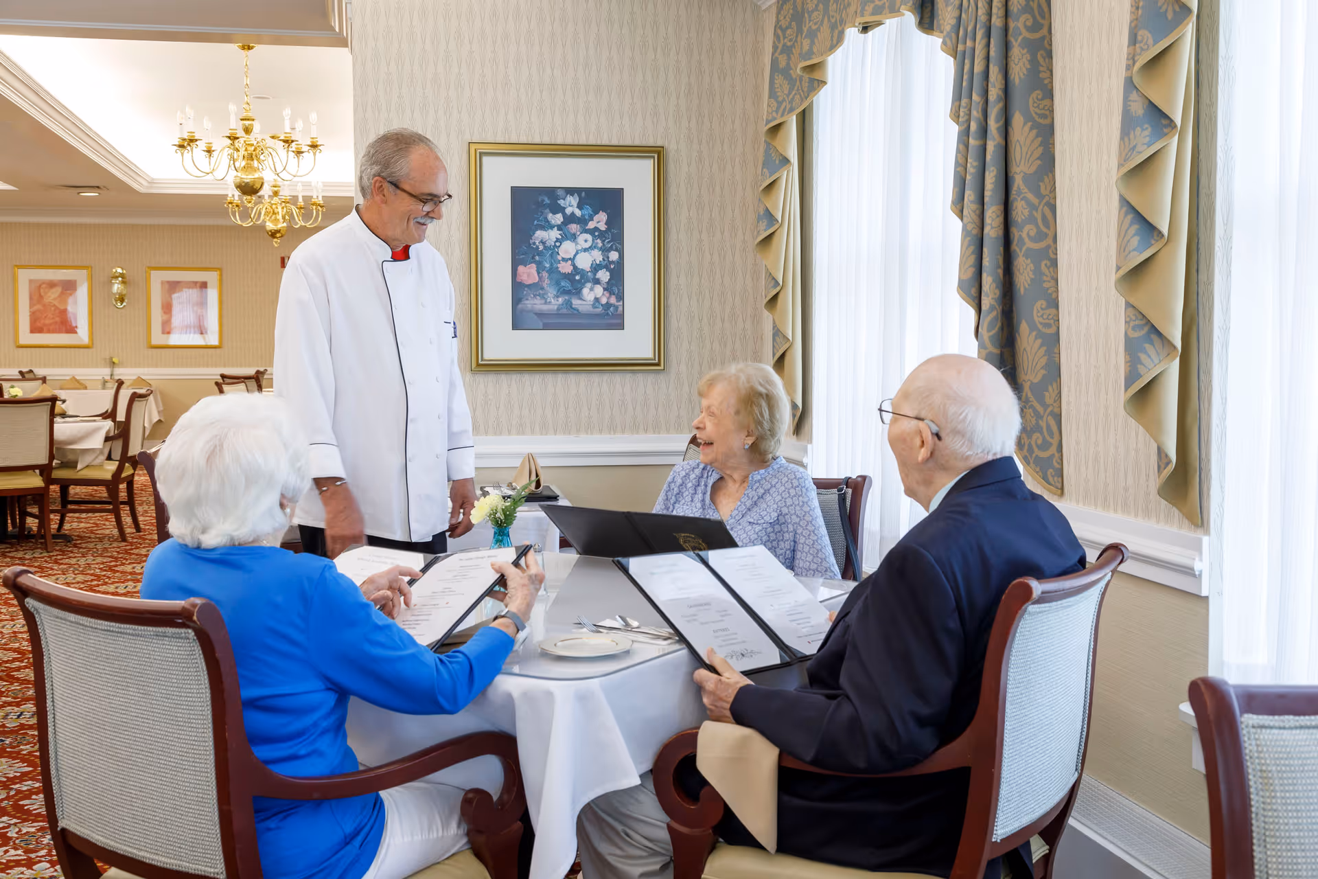 Three elderly people sitting at a round dining table with menus in hand, interacting with a chef in a white uniform in an elegant dining room with patterned wallpaper, framed artwork, and large windows with draped curtains.