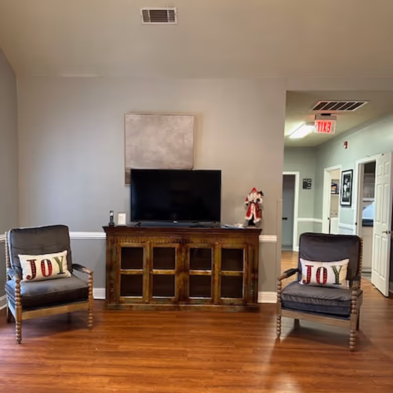 A living room area with a TV on a wooden console flanked by two armchairs each with 'JOY' pillows.
